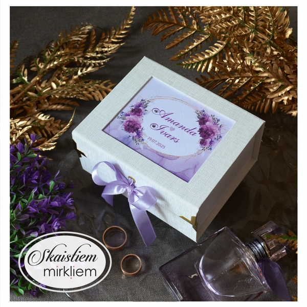 Balta gredzenu kastīte ar violetiem ziediem un vārdiem "Amanda un Ivars"