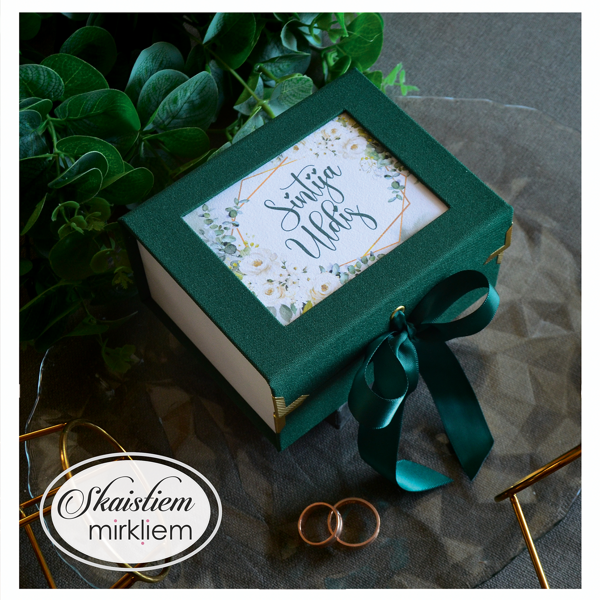 Tumši zaļa gredzenu kastīte ar ziedu rāmi un vārdiem "Sintija un Uldis"