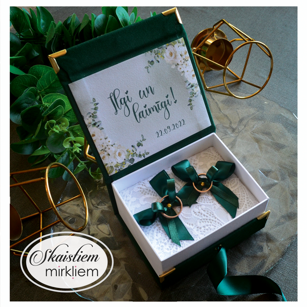 Tumši zaļās gredzenu kastītes atvērums ar ziedu rāmi un vārdiem "Sintija un Uldis"