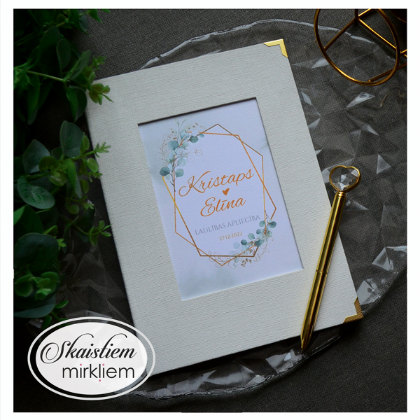 Pienbalti laulības apliecības vāki ar zelta sešstūra rāmi un vārdiem "Kristaps un Elīna"