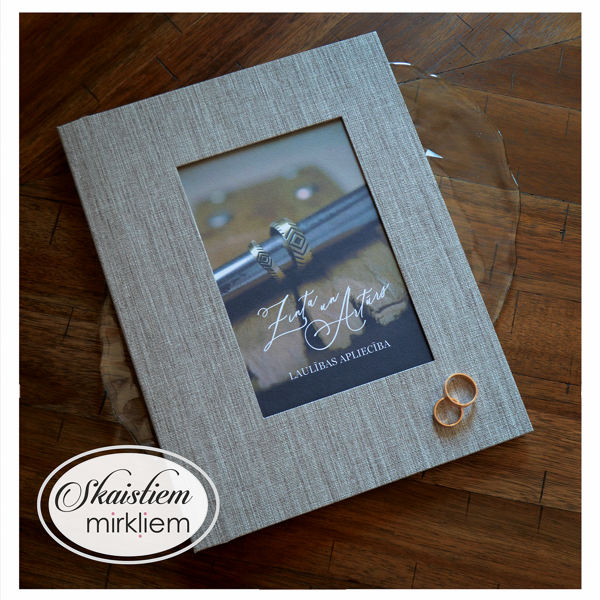 Lina tekstūras laulības apliecības vāki ar tumšu fonu, kur ir nodrukāti viņu gredzeni