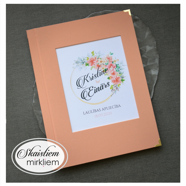 Persiku krāsas laulības apliecības vāki ar ziedu vainagu un vārdiem "Kristīne un Einārs"