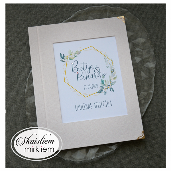 Gaiši rozā laulības apliecības vāki ar zelta sešstūra rāmi un vārdiem "Betija un Rihards"