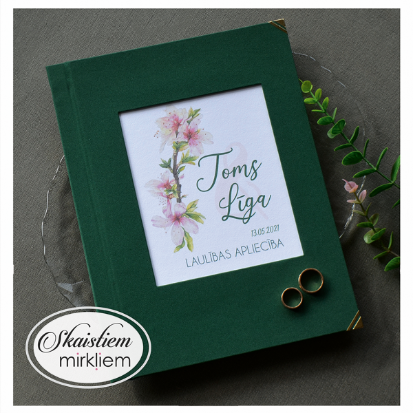 Tumši zaļi laulības apliecības vāki ar ābeļziedu akcentu un vārdiem "Toms un Līga"