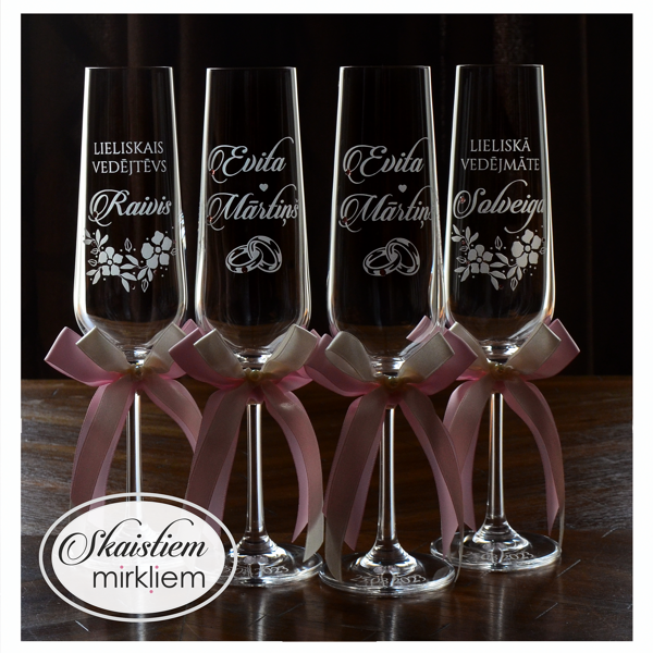 Kāzu glāzes ar vārdu, gredzenu un ziedu gravējumu, kam pieskaņotas rozā lentītes